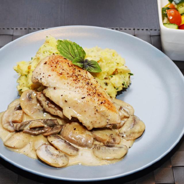 sur une assiette blanche posée sur un set tressé, un filet de volaille avec sa sauce aux champignons, accompagné d'un peu de gratin dauphinois. Dans le fond de l'image, on aperçoit un bol avec une salade composée.