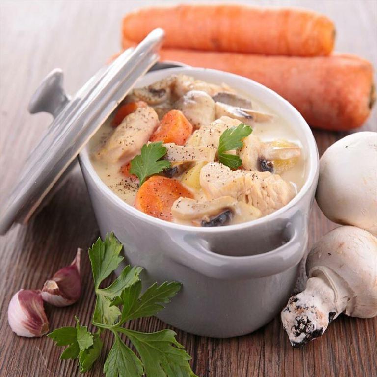 Une terrine ovale en porcelaine blanche, le couvercle ouvert emplie de blanquette de veau ; des carottes, des champignons, de l'al forment le décor de la photo
