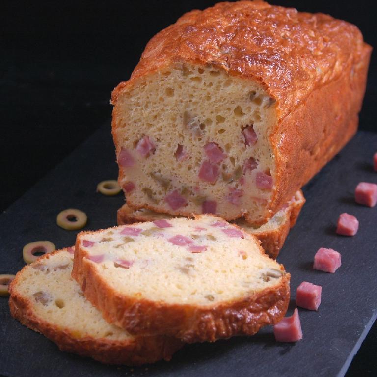 sur une plateau en ardoise grise, un cake aux olives et au dés de jambon, avec trois tranches découpées, le restant du cake et des rondelles d'olive et de petits dés de jambon en décoration