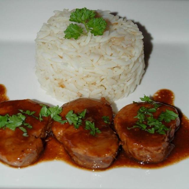 sur une assiette blanche, trois médaillons de filet mignon de porc, nappés d'une sauce brun foncé ; en arrière plan, du riz présenté sous forme de cylindre, quelques feuilles de cerfeuil en décoration