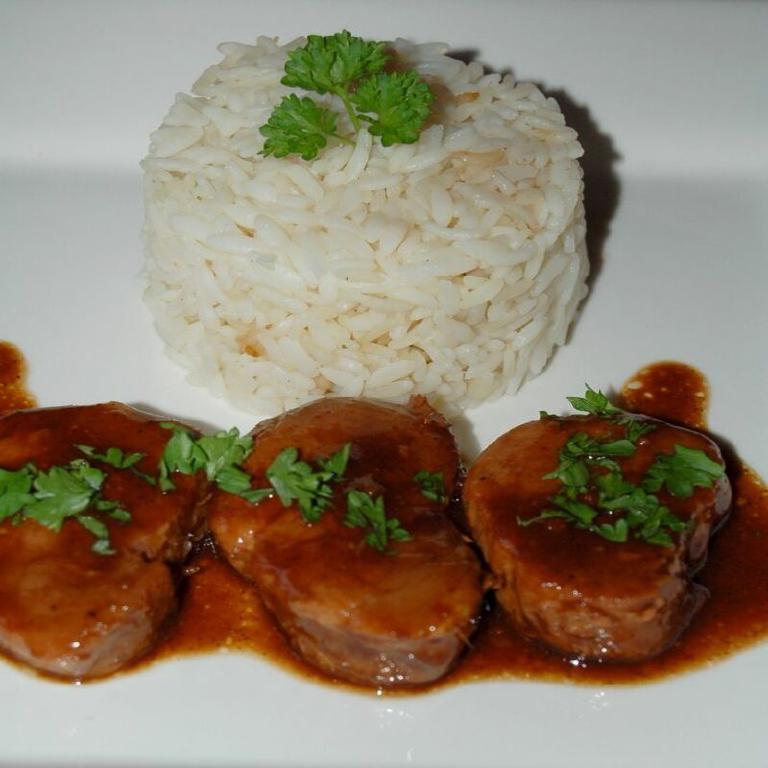 sur une assiette blanche, trois médaillons de filet mignon de porc, nappés d'une sauce brun foncé ; en arrière plan, du riz présenté sous forme de cylindre, quelques feuilles de cerfeuil en décoration