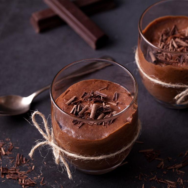 une verrine emplie de mousse au chocolat ; la verrine est entourée d'un morceau de ficelle ; quelques copeaux de chocolat parsèment la mousse. Dans le fond, une autre verrine, une cuillère, des bâtonnets de chocolat