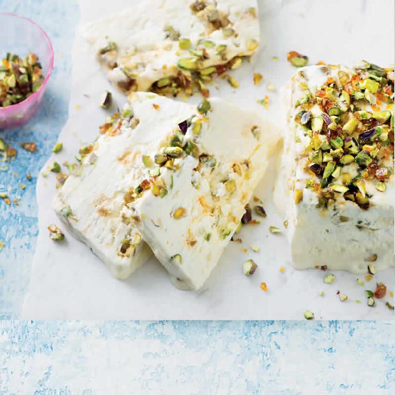 sur un plat en faïence blanceh, trois tranches de nougat glacé et à côté le restant du nougat, recouvert de brisures de pistaches et d'amandes. Les teintes de la photo sont dans le blanc, le vert, le jaune.