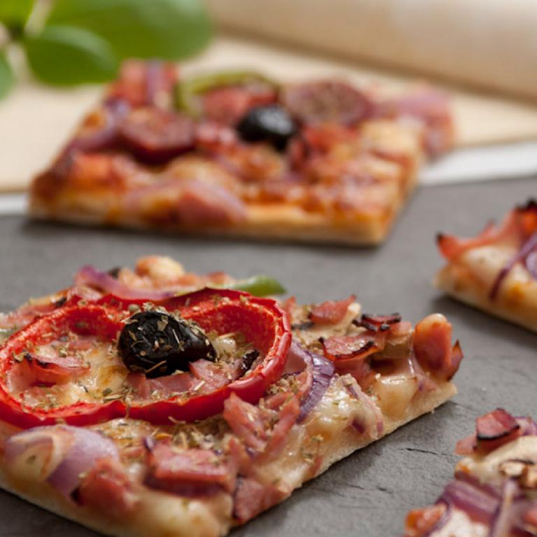un morceau de pizza reposant sur une plaque à pizza, avec dans le fond d'autres morceaux, vus de manière floue. La pizza est couverte d'olives, de dés de jambon, de tomates fraîches, d'herbe séchées