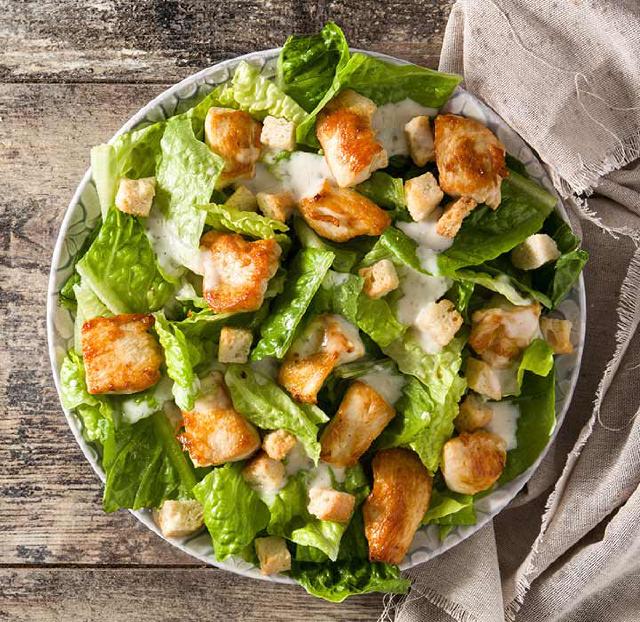 sur une table en bois, une assiette creuse avec une salade césar, des morceaux de blancs de poulet, des morceaux de salades, quelques dés de pain grillé. L'assiette est en partie posée sur un torchon