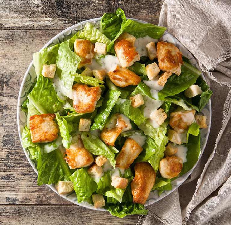 sur une table en bois, une assiette creuse avec une salade césar, des morceaux de blancs de poulet, des morceaux de salades, quelques dés de pain grillé. L'assiette est en partie posée sur un torchon