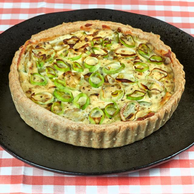 sur un plat rond en céramique sombre, une tarte aux poireaux et à la ricotta, décorée avec des rondelles d'oignons nouveaux