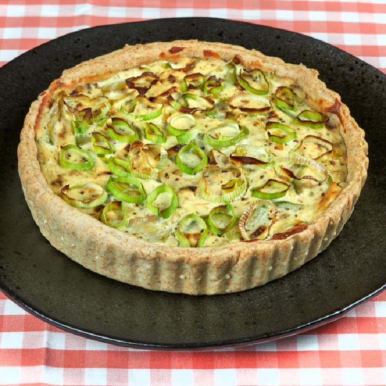 sur un plat rond en céramique sombre, une tarte aux poireaux et à la ricotta, décorée avec des rondelles d'oignons nouveaux