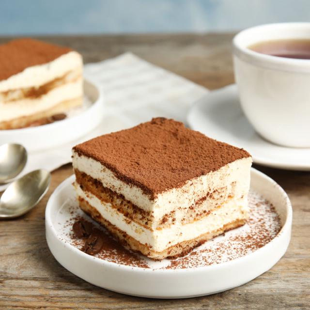 une assiette avec un morceau de tiramisu recouvert de poudre de cacao ; sur la table, on aperçoit une tasse à thé et un autre morceau de tiramisu