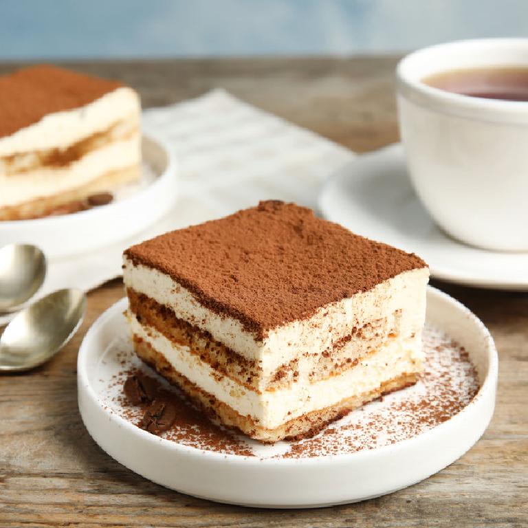 une assiette avec un morceau de tiramisu recouvert de poudre de cacao ; sur la table, on aperçoit une tasse à thé et un autre morceau de tiramisu